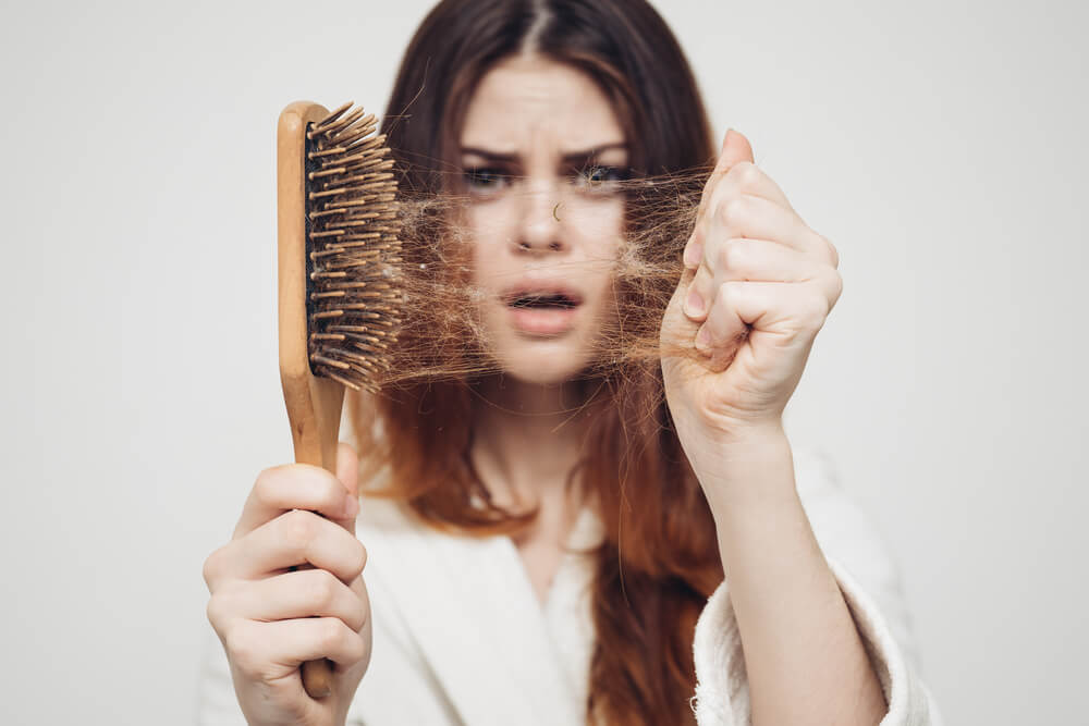 Na foto uma moça com cabelos longos segurando uma escova cheia de  cabelos com cara de espanto