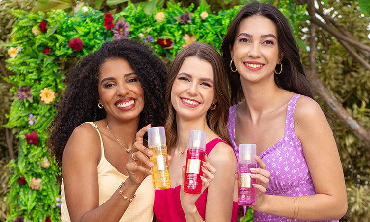 Três mulheres sorrindo segurando frascos de body splash juntas em frente a folhagem e flores