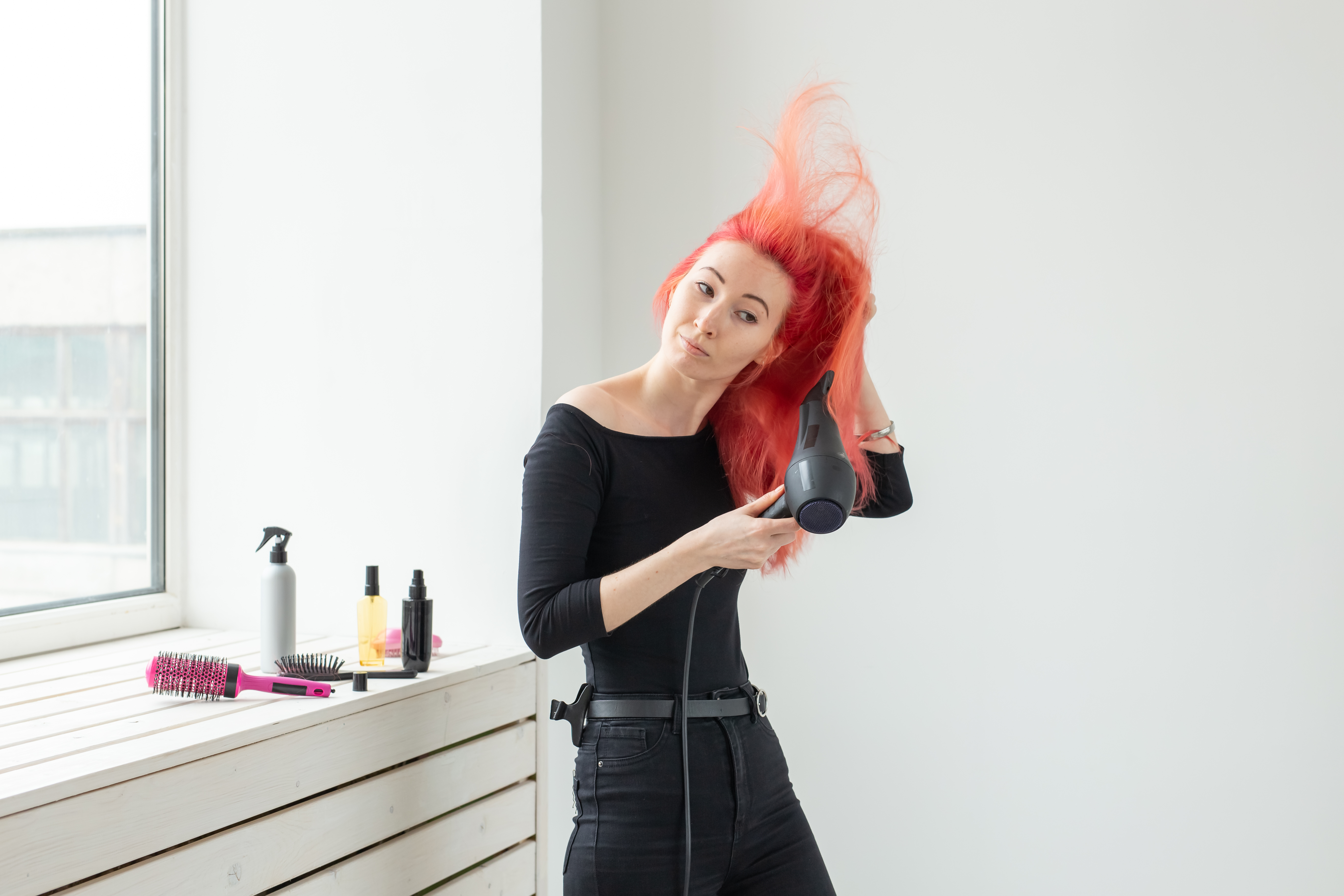 Mulher secando os fios em casa, destacando cuidados com cabelos coloridos em cabelo vermelho vibrante, com secador e produtos capilares ao lado.