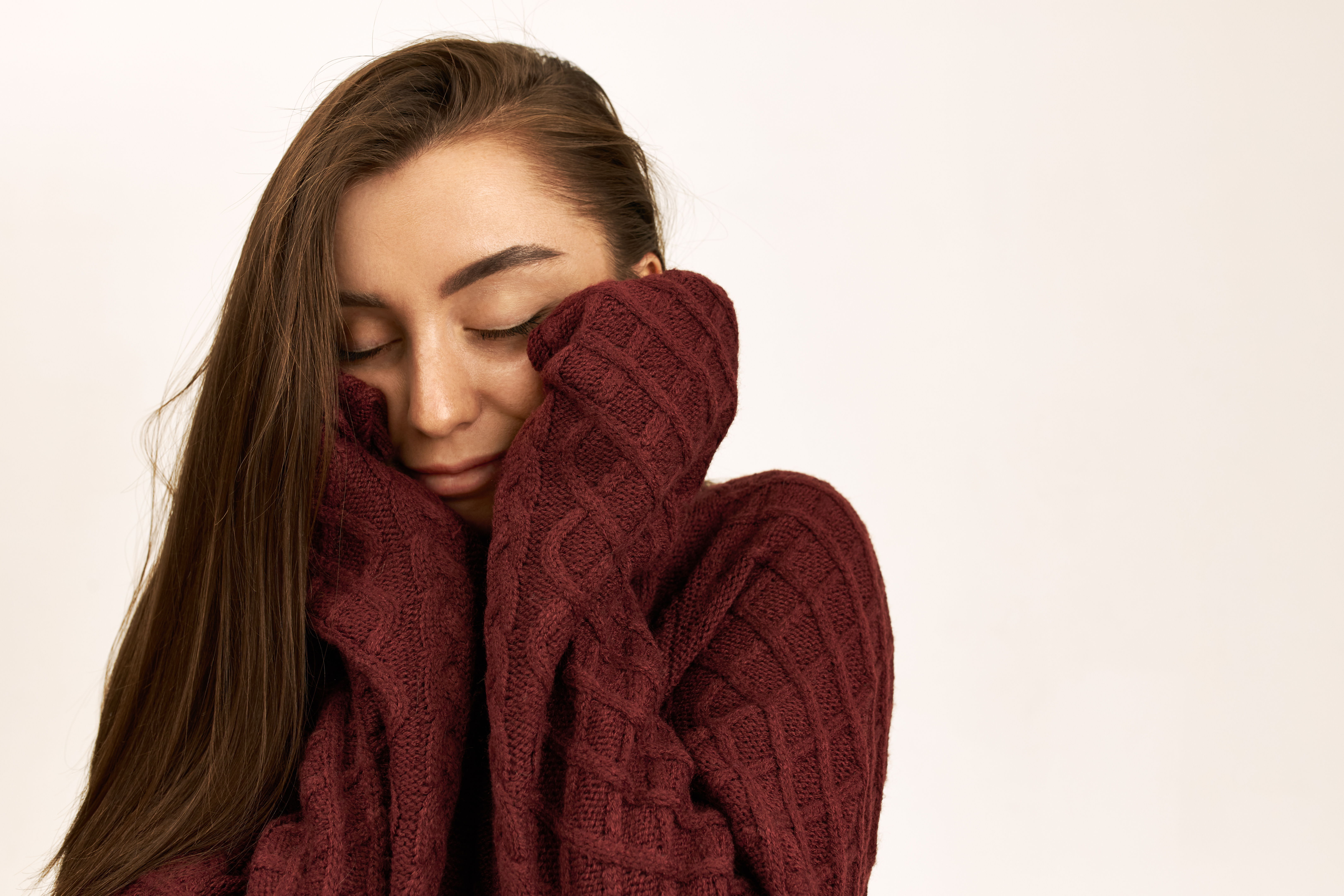 Jovem sorridente com os olhos fechados e cabelos castanhos soltos, vestindo um suéter de lã vinho que remete aos cuidados com o cabelo no inverno contra o ressecamento e o frizz.